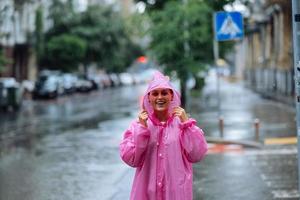 ung leende kvinna med regnkappa medan njuter en regnig dag. foto