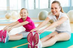 sportig mor och dotter. glad mor och dotter håller på med stretching övningar och leende medan Sammanträde på övning mattor i sporter klubb foto