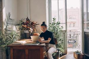 koncentrerad ung kvinna använder sig av digital läsplatta medan Sammanträde på henne arbetssätt plats i kontor foto