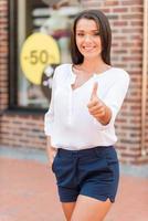 bra försäljning skön ung kvinna som visar henne tumme upp och leende medan stående mot Kläder Lagra foto