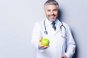 äta friska glad mogna läkare i ger en grön äpple till du och leende medan stående mot grå bakgrund foto