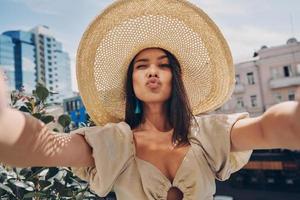 lekfull ung kvinna i elegant hatt framställning selfie medan avkopplande på de balkong foto