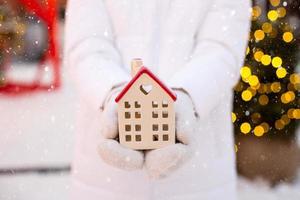 hus, miniatyr- stuga i händer av kvinna bär vantar och värma kläder utomhus- i snö. handla för verklig egendom, inköp, konstruktion, flytt, inteckning. mysigt Hem, jul, ny år bokning foto