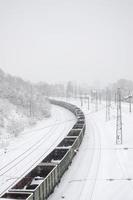 en lång tåg av frakt bilar är rör på sig längs de järnväg Spår. järnväg landskap i vinter- efter snöfall foto