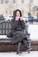 en ung kvinna varmt klädd är Sammanträde på en bänk i de stad på de gata på en snöig kall dag foto