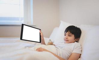Lycklig unge liggande i säng innehav läsplatta tittar på tecknad serie och chattar med vänner på digital vaddera, söt ung pojke som visar Rör vaddera, barn avkopplande i de morgon- innan gå till skola foto