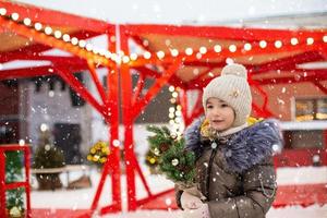 en flicka med en jul träd i henne händer utomhus- i värma kläder i vinter- på en festlig marknadsföra. fe- lampor kransar dekorerad snö stad för de ny år foto