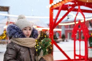 en flicka med en jul träd i henne händer utomhus- i värma kläder i vinter- på en festlig marknadsföra. fe- lampor kransar dekorerad snö stad för de ny år foto
