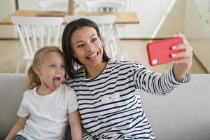 mamma, liten dotter ta selfie Foto förbi telefon, visa tungor tillsammans, har roligt, Sammanträde på soffa