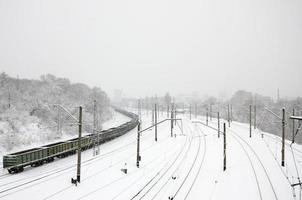 en lång tåg av frakt bilar är rör på sig längs de järnväg Spår. järnväg landskap i vinter- efter snöfall foto