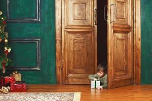 barn pojke kikar ut av de dörr med en jul gåva låda. foto