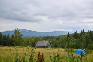landskap, grön äng, blå tält, trä- hus, fält gräs, skön se av de berg toppar i de moln. foto