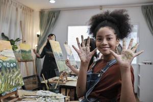 afrikansk amerikan flicka visar henne hand trasslat upp med akryl färger, ler och utseende på kamera, roligt lär med studerande barn i konst studio klass, kreativ målning med Kompetens i skola utbildning. foto