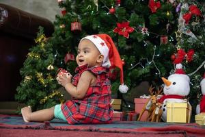 afrikansk amerikansk baby ler lyckligt medan den tar emot liten presentask från föräldrar medan han klär sig i julklänning och tomtehatt med julgran på baksidan för säsongsfirande koncept foto