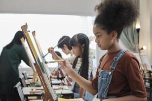 afrikansk amerikan flicka koncentrat på akryl Färg bild målning på duk med studenter grupp i konst klassrum, kreativ inlärning med talanger och Kompetens i elementärt skola studio utbildning. foto