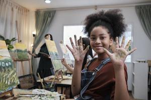 afrikansk amerikan flicka visar henne hand trasslat upp med akryl färger, ler och utseende på kamera, roligt lär med studerande barn i konst studio klass, kreativ målning med Kompetens i skola utbildning. foto