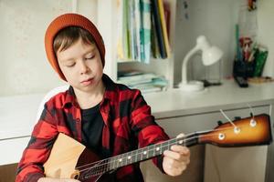 pojke i en röd hatt och en pläd skjorta spelar de balalajka. stilig pojke innehav hans gitarr. musik lektioner på Hem. hobby för de själ. Hem undervisning musik foto