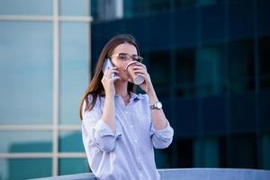 verkställande företag kvinna talande på mobil smartphone och dricka kaffe från disponibel papper kopp i de gata med kontor byggnader i de bakgrund foto