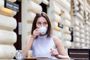 skön asiatisk ung kvinna med kopp av kaffe. kvinna åtnjuter färsk kaffe i de morgon- med soluppgång på coffe affär skön kvinna dricka kaffe i de morgon- Sammanträde utomhus foto