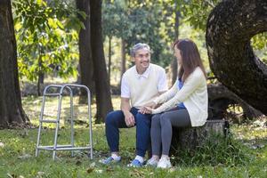 asiatisk senior far med rollator och dotter Sammanträde tillsammans under de stor träd i de parkera under sommar för ljus övning och fysisk terapi begrepp foto