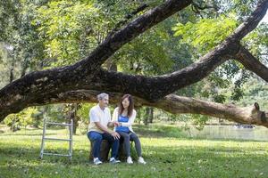 asiatisk senior far med rollator och dotter Sammanträde tillsammans under de stor träd i de parkera under sommar för ljus övning och fysisk terapi begrepp foto