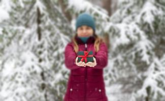 ung kvinna i defokusering i röd jacka innehav i händer gåva grön låda med röd band mot snöig gran träd i vinter- skog jul och ny år högtider närvarande foto