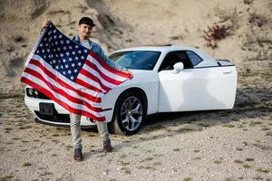 stilig man i jeans jacka och keps med USA flagga nära hans vit amerikan muskel bil i karriär. foto