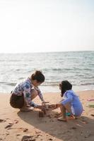 asiatisk söt liten flicka och henne mor spelar eller framställning sand slott eller grävning med sand på tropisk strand. barn med skön hav, sand blå himmel. Lycklig barn på semester havet på de strand. foto
