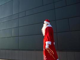 en ung man klädd som santa på de gata, stående mot en grå modern vägg. vit skägg, en röd kostym med en hatt. jul, ny år. ryska farfar glasera. grå modern bakgrund, kopia utrymmen foto