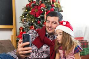 pappa och dotter nära de jul träd med en smartphone ta en selfie, kommunicera via video förbindelse. jul hälsningar, en gåva låda, en flicka i en santa hatt vinka Hallå. ny år, Semester dekor foto