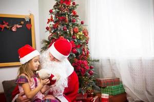 liten flicka i santa hatt på jultomten knä nära jul träd i jul dekor. visar en låda med en gåva, en mjuk leksak, finputsning hans skägg och skrattar. ny år, ryska farfar frost foto