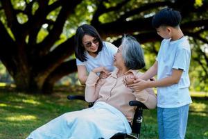 glad senior asiatisk mormor använder rullstol med sin dotter och barnbarn i parken, barnbarn kom för att besöka äldre mormor och hålla handen. begreppet lycklig familj, bra relation tillsammans foto