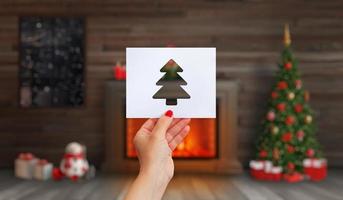 hand innehar en papper med en jul träd skära ut på Det. öppen spis med en brand, en jul träd och jul dekorationer foto