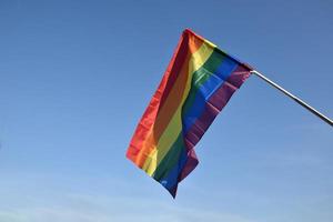 regnbåge flagga innehav i hand mot blå himmel bakgrund, begrepp för HBTQ firande i stolthet månad, juni, runt om de värld. foto