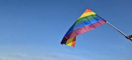 regnbåge flagga innehav i hand mot blå himmel bakgrund, begrepp för HBTQ firande i stolthet månad, juni, runt om de värld. foto