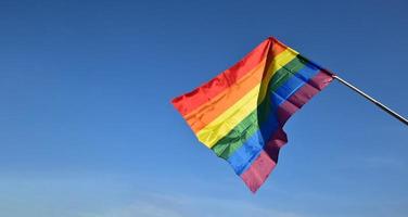 regnbåge flagga innehav i hand mot blå himmel bakgrund, begrepp för HBTQ firande i stolthet månad, juni, runt om de värld. foto