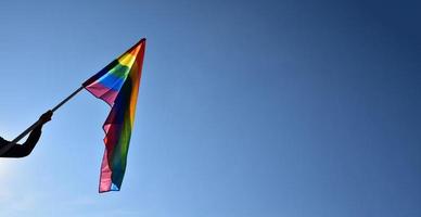 regnbåge flagga innehav i hand mot blå himmel bakgrund, begrepp för HBTQ firande i stolthet månad, juni, runt om de värld. foto