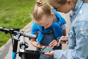 mor och dotter använder sig av Ansökan på smartphone för delning elektrisk skotrar foto