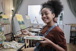 ett svart studerande flicka leende med paintbrush och palett, akryl Färg bild målning på duk i konst klassrum och kreativ inlärning med talanger skicklighet på elementärt skola studio utbildning. foto