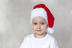 porträtt av en söt liten pojke i en vit t-shirt och santa claus hatt. barns känslor. jul och ny år. Framgång, ljus aning, kreativ idéer och begrepp. foto