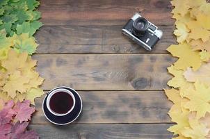 en kopp av te och ett gammal kamera bland en uppsättning av gulning fallen höst löv på en bakgrund yta av naturlig trä- styrelser av mörk brun Färg foto