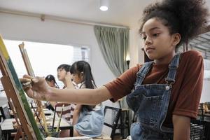 afrikansk amerikan flicka koncentrat på akryl Färg bild målning på duk med studenter grupp i konst klassrum, kreativ inlärning med talanger och Kompetens i elementärt skola studio utbildning. foto