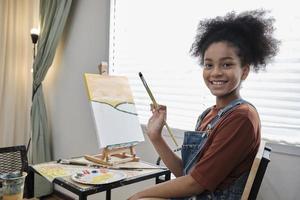 ett afrikansk amerikan flicka ser på kamera och leende, akryl Färg bild målning på duk i konst klassrum och kreativ inlärning med talanger och skicklighet på elementärt skola studio utbildning. foto