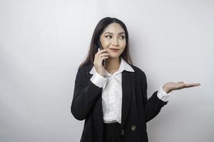 upphetsad asiatisk affärskvinna bär svart kostym pekande på de kopia Plats bredvid henne medan talande på henne telefon, isolerat förbi vit bakgrund foto
