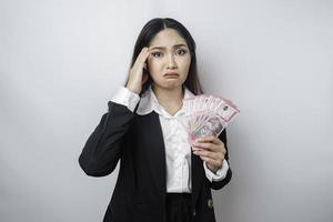en förvirrad ung affärskvinna är bär svart kostym och innehav kontanter pengar i indonesiska rupiah isolerat förbi vit bakgrund foto