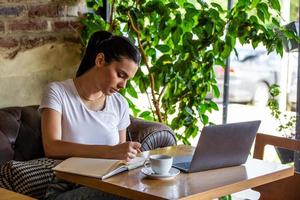 ung kvinna med kaffe kopp och bärbar dator på coffe affär. kvinna tar en ha sönder. njuter arbete från kaffe affär. håller på med företag från kaffe affär foto