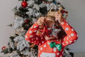 liten lockigt hår flicka i en karneval klänning gömde sig henne ansikte Bakom skinande jul träd leksaker stjärnor. barn i en röd klänning med en santa skriva ut på de bakgrund av en jul träd. hög kvalitet Foto