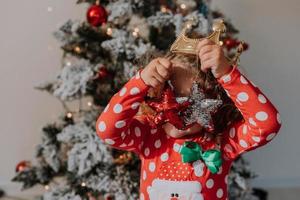 liten lockigt hår flicka i en karneval klänning gömde sig henne ansikte Bakom skinande jul träd leksaker stjärnor. barn i en röd klänning med en santa skriva ut på de bakgrund av en jul träd. hög kvalitet Foto