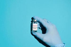 hand i blå medicinsk handskar innehav en injektionsflaska av covid-19 vaccin på en blå bakgrund, topp se. vaccin produktion begrepp, global immunisering av de befolkning. foto