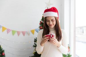 leende brunett kvinna i Tröja och santa hatt innehav små gåva låda och gläds över jul dekorativ vit bakgrund foto